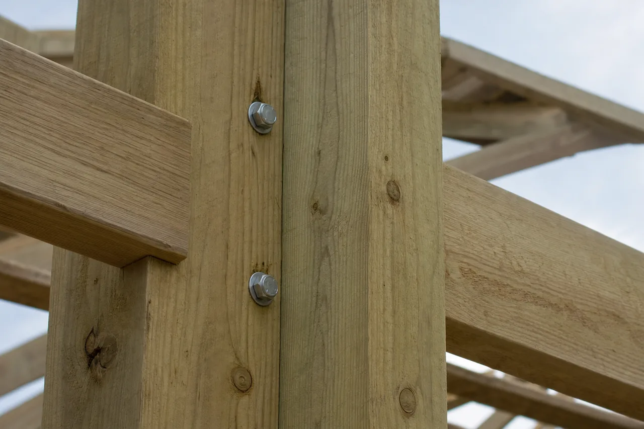 Two square timber posts meeting horizontal girts at a corner with galvanized bolts against a pale sky — pole barn, Tyler. Illustrative example.